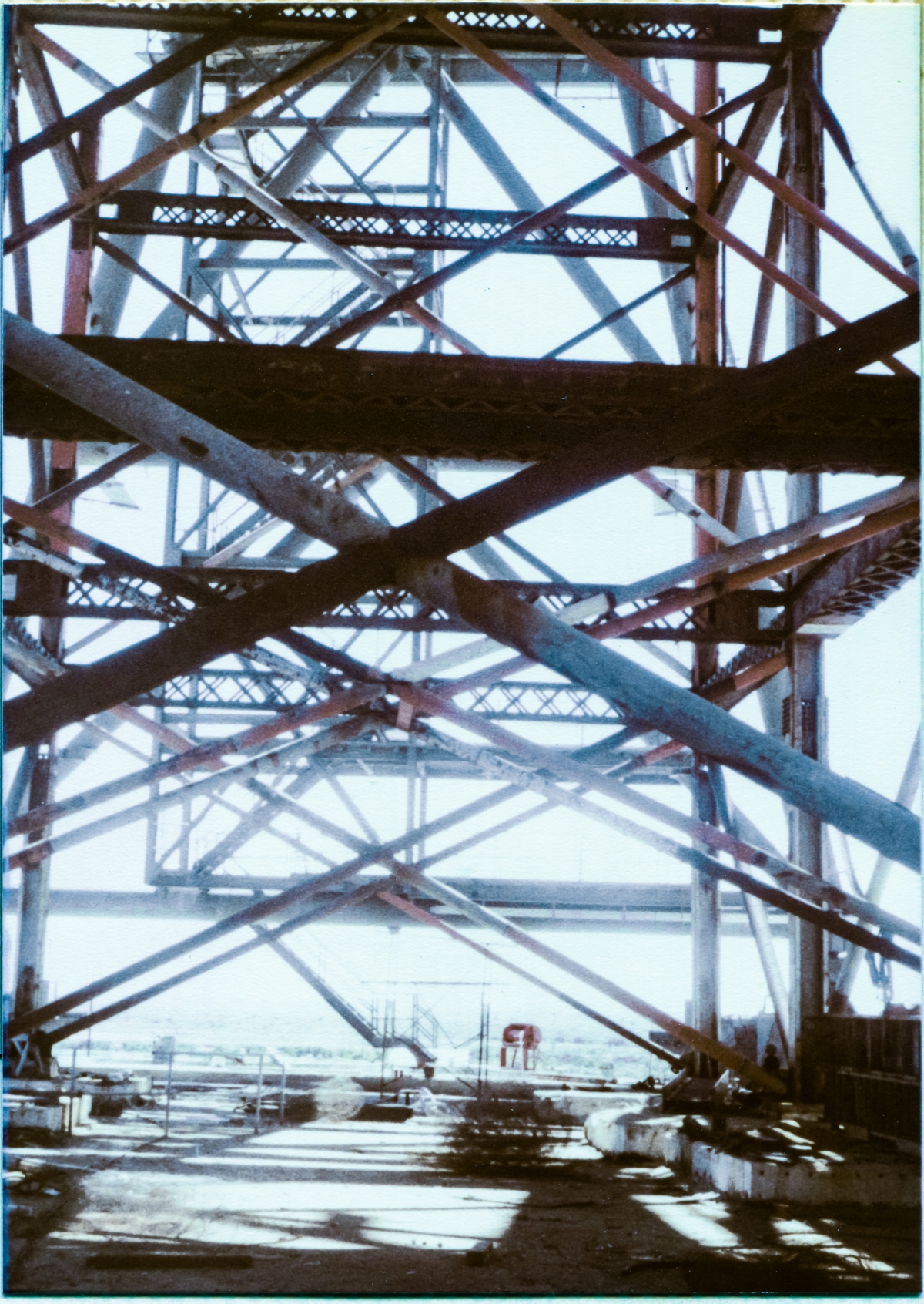 Image 016. Viewed from a vantage point on the Pad Deck, immediately south of the Fixed Service Structure at Space Shuttle Launch Complex 39-B, Kennedy Space Center, Florida, the Falsework which is holding up the Rotating Service Structure presents itself as a confusing maze of diagonal pipe bracing, tying heavy vertical and horizontal Falsework framing members together. The Falsework was erected on the Pad Deck, resting on temporary concrete footings, before the RSS was then assembled on top of it, and it held the growing RSS up in the air until it became complete enough to support itself. Farthest from the camera, the heavy structural pipes of RSS Column Line 7 with the offset steel framing of Stair Tower 3 straddling them, extending from the ground all the way up to elevation 135’-7” which is the main floor elevation, define the far end of the RSS, which you are standing directly beneath, as seen in this view. Photo by James MacLaren.